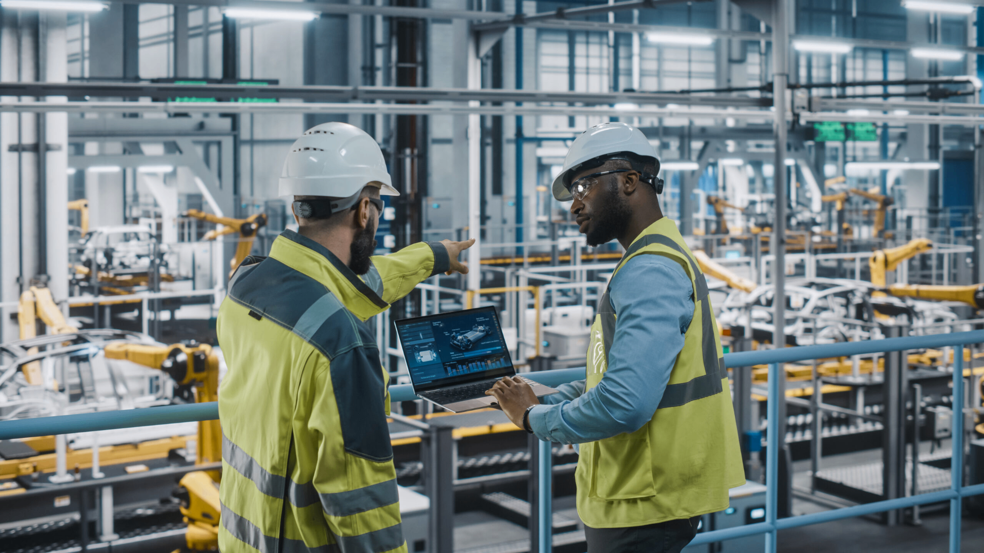 Factory workers with safety equipment examining manufacturing operations on laptop